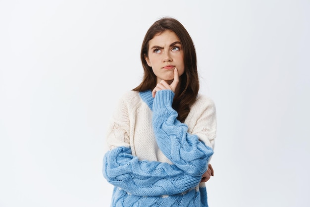 Thoughtful girl staring at upper left corner and frowning serious thinking and standing focused in sweater making plan or searching solution white background