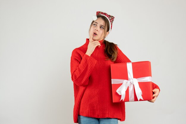 thoughtful girl with santa hat holding present standing on white