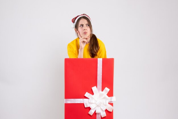 thoughtful girl with santa hat standing behind big xmas gift on white