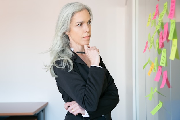 Thoughtful gray-haired businesswoman reading notes on glass wall and holding marker. Concentrated Caucasian female worker in suit thinking about idea for project. 