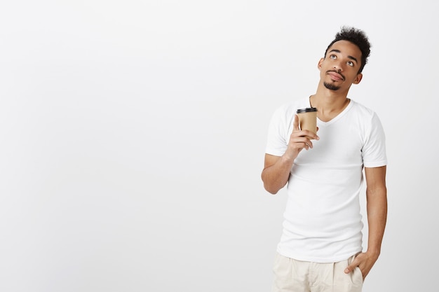 Thoughtful handsome Black man in casual t-shirt looking dreamy up and drinking coffee