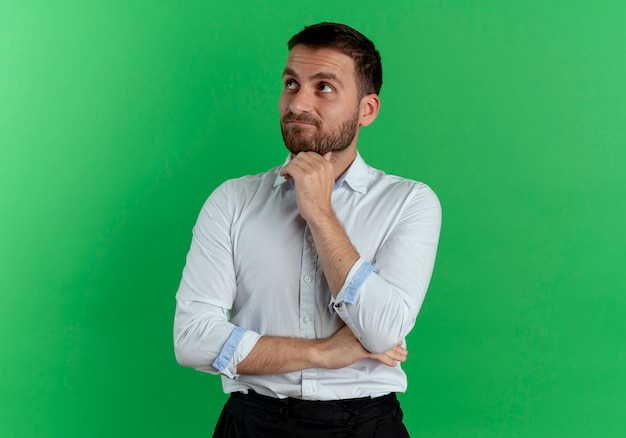 Thoughtful handsome man puts hand on chin looking up isolated on green wall