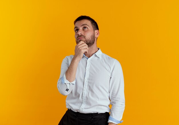 Thoughtful handsome man puts hand on chin looking up isolated on orange wall