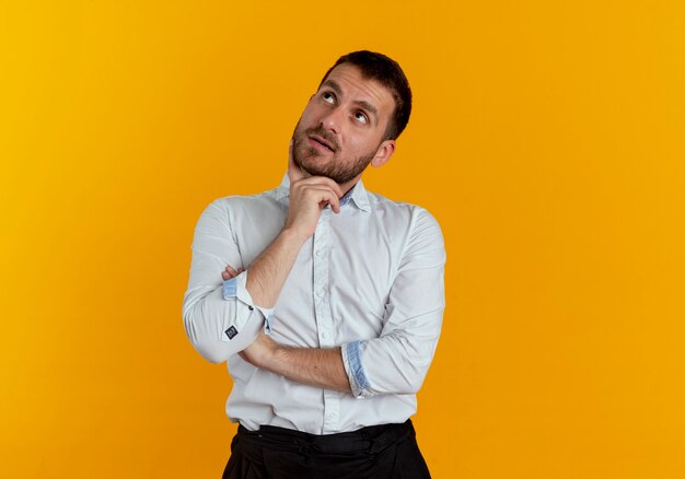 Thoughtful handsome man puts hand on chin looking up isolated on orange wall