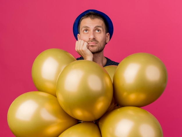 Thoughtful handsome man wearing blue party hat puts hand on chin and holds helium balloons isolated on pink wall with copy space