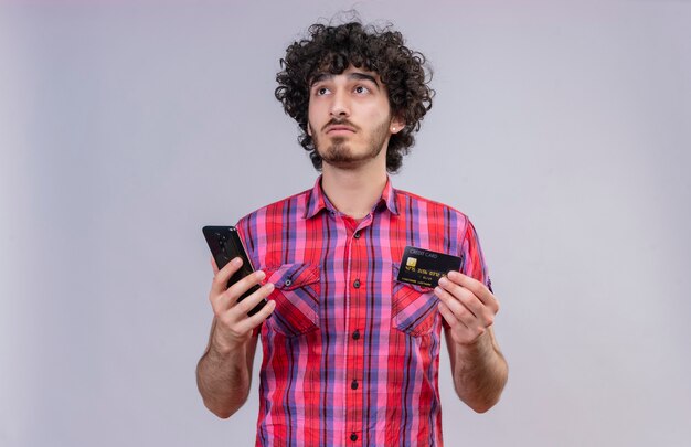 Вдумчивый красавец с вьющимися волосами в клетчатой рубашке с кредитной картой и мобильным телефоном