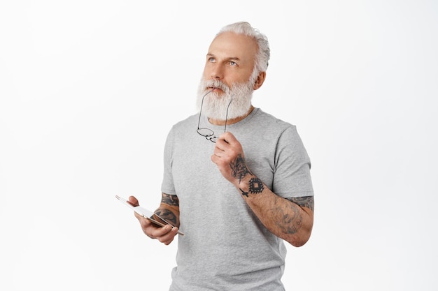 Free photo thoughtful hipster senior man biting temple of glasses, looking up pondering while using digital tablet, shopping online, standing against white background