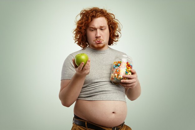 вдумчивый нерешительный рыжий толстый мужчина с большим животом, чувствующий себя смущенным и нерешительным, сталкивается с трудным выбором: есть ли здоровое органическое яблоко или нездоровые сладости