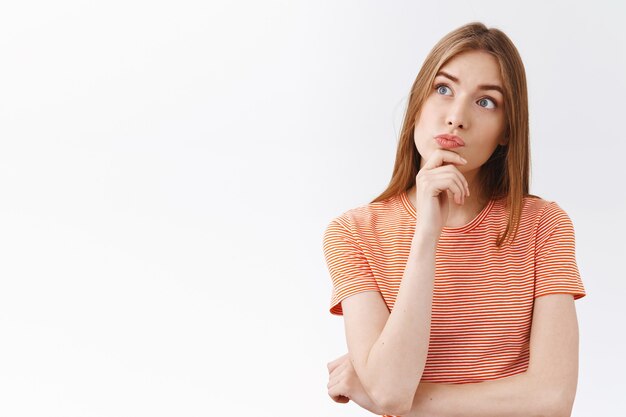 Thoughtful intrigued young fair-haired girl in striped t-shirt, touch chin and pouting curious, look upper left corner intrigued and unsure, making choice, thinking about decision, white background