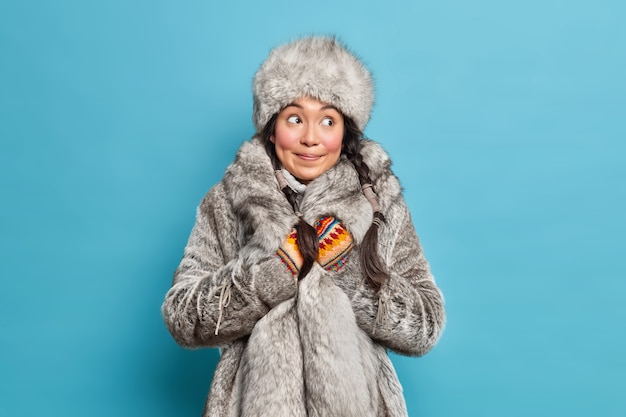 Задумчивая инуитская женщина, одетая в серую меховую шапку и пальто, вязаные варежки, носит теплую зимнюю одежду, изолированную над синей стеной студии