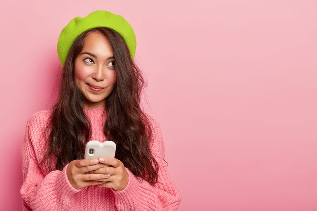 Thoughtful Korean teenage girl with long dark hair, uses mobile phone for messaging online, wears green beret and knitted sweater