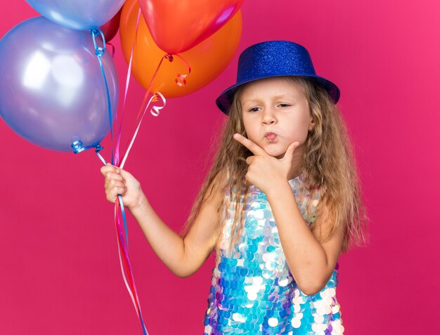 thoughtful little blonde girl with blue party hat putting hand on chin and holding helium balloons isolated on pink wall with copy space