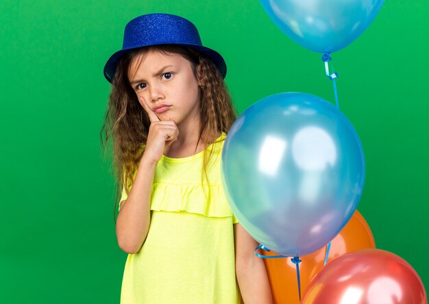 턱에 손가락을 넣고 복사 공간이 녹색 벽에 고립 된 헬륨 풍선을 들고 파란색 파티 모자와 사려 깊은 어린 백인 소녀