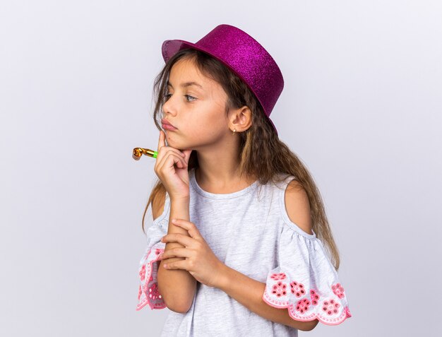 thoughtful little caucasian girl with purple party hat putting hand on chin holding party whistle and looking at side isolated on white wall with copy space