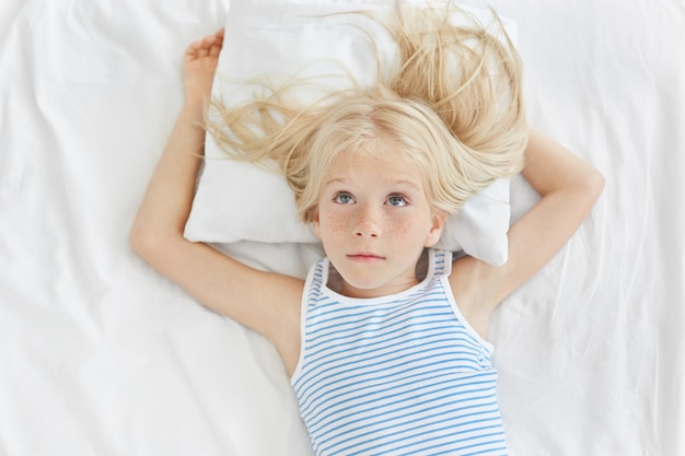 Bambina premurosa con lunghi capelli biondi, alzando lo sguardo, indossa camicia a righe, sdraiato sul cuscino bianco, sognando nuova bicicletta. ragazza adorabile riposante che riposa sul letto bianco nella stanza del `s dei bambini