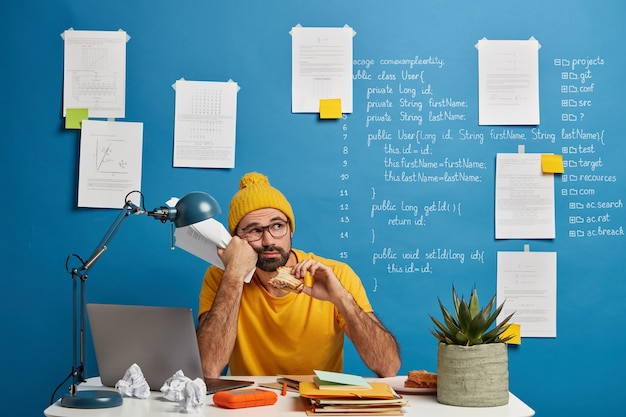 Thoughtful male programmer or software developer ponders on program code, looks away and eats burger, holds papers wears yellow clothes spends time for making project.