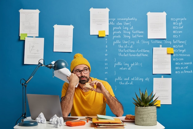Вдумчивый мужчина-программист или разработчик программного обеспечения размышляет над программным кодом, смотрит в сторону и ест бургер, держит бумаги, носит желтую одежду, тратит время на создание проекта.