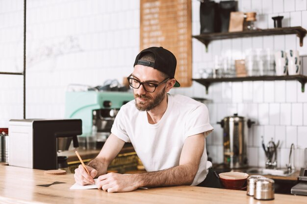 Thoughtful man in eyeglasses and cap standing behind bar counter and writing in notepad while working in cafe