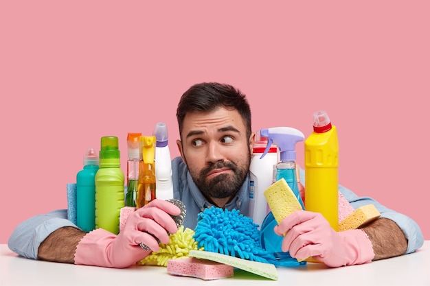 Foto gratuita l'uomo premuroso si sente oberato di lavoro con le pulizie domestiche, guarda pensieroso da parte, si siede alla scrivania con prodotti chimici