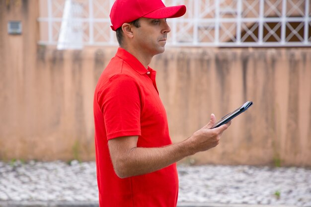 Заботливый мужчина держит таблетку, стоя и глядя в сторону. Обрезанный вид сбоку кавказского курьера в красной форме, ищущего нужный адрес. Выборочный фокус. Служба доставки и почтовая концепция