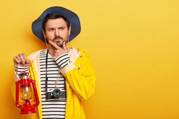 Thoughtful man holds chin, thinks about having journey or expedition, holds little gas lamp, dressed in raincoat, headgear, uses camera for making photos, isolated over yellow wall