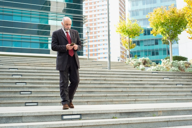 Вдумчивый зрелый человек с помощью телефона во время спуска по лестнице