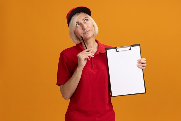 Thoughtful middle-aged blonde delivery woman in red uniform and cap holding clipboard touching face with pencil looking up isolated on orange wall with copy space