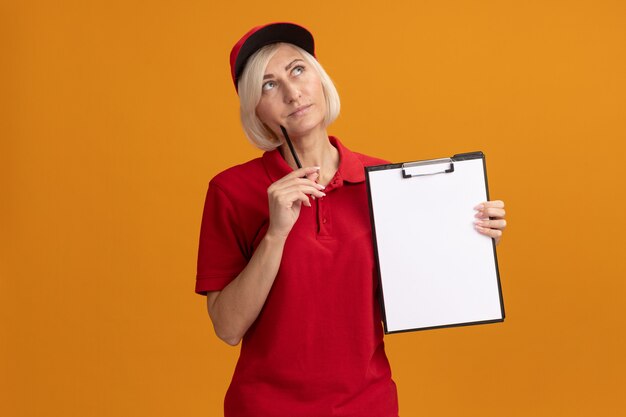 赤い制服を着た思いやりのある中年の金髪の配達の女性とコピースペースでオレンジ色の壁に分離された鉛筆で顔に触れるクリップボードを保持しているキャップ