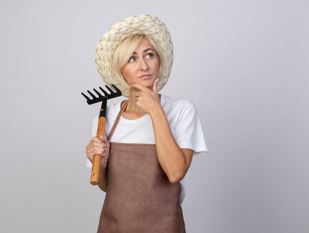 Thoughtful middle-aged blonde gardener woman in uniform wearing hat holding rake keeping hand on chin looking at side isolated on white wall with copy space