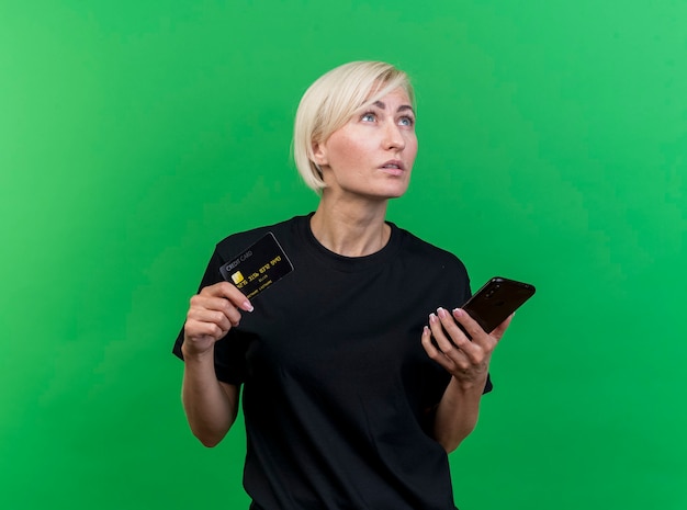 Thoughtful middle-aged blonde slavic woman holding credit card and mobile phone looking up isolated on green background with copy space