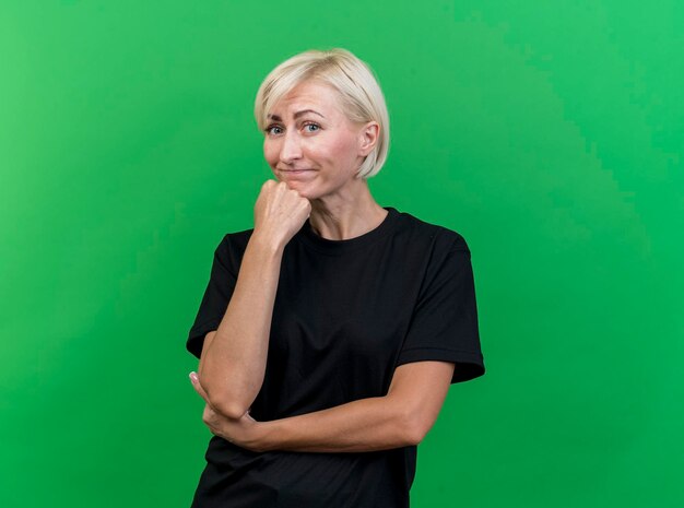 Thoughtful middle-aged blonde slavic woman putting hand on chin looking at front isolated on green wall with copy space