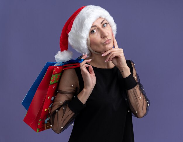 thoughtful middle-aged blonde woman wearing christmas hat holding christmas gift bags on shoulder looking  touching face isolated on purple wall