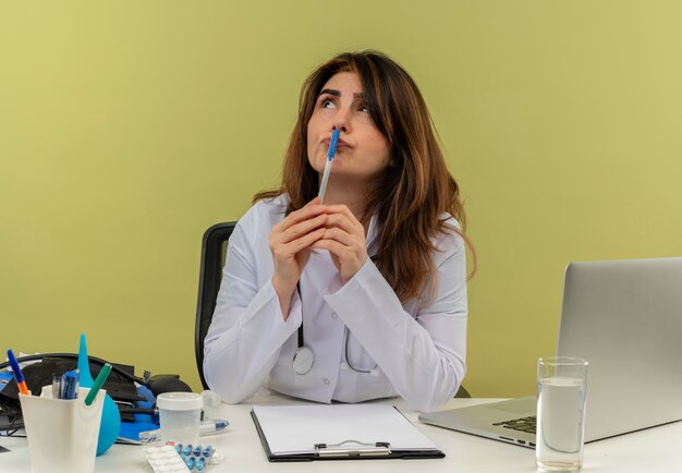 Thoughtful middle-aged female doctor wearing medical robe and stethoscope sitting at desk with medical tools clipboard and laptop looking up touching lips with pen isolated
