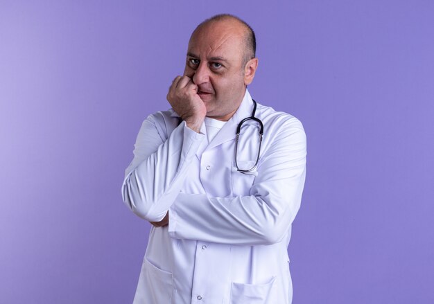 thoughtful middle-aged male doctor wearing medical robe and stethoscope keeping hand on chin looking at front isolated on purple wall with copy space