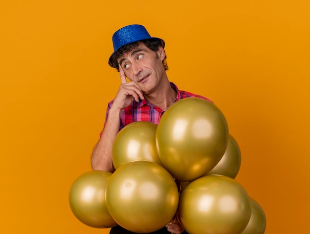 Uomo di mezza età premuroso che indossa il cappello del partito in piedi dietro i palloncini che mette il dito sul tempio che guarda il lato isolato sulla parete arancione con lo spazio della copia