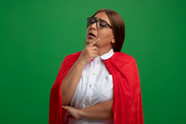 thoughtful middle-aged superhero female looking at side wearing glasses putting hand under chin isolated on green