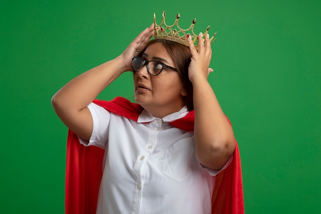 thoughtful middle-aged superhero female looking up putting crown on head isolated on green