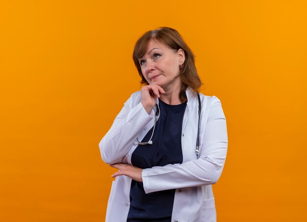 Thoughtful middle-aged woman doctor wearing medical robe and stethoscope putting handon chin on isolated orange wall with copy space