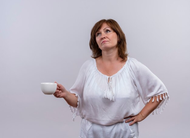 Thoughtful middle-aged woman holding cup of tea looking up on isolated white wall