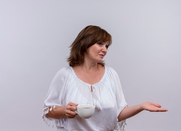 Thoughtful middle-aged woman holding cup of tea and putting hand to side on isolated white wall