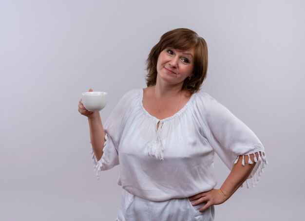 Thoughtful middle-aged woman holding cup of tea and putting hand on waist on isolated white wall