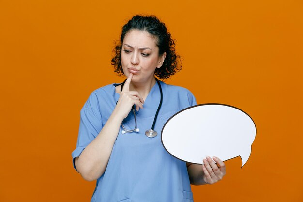 Thoughtful middleaged female doctor wearing uniform and stethoscope around her neck holding chat bubble touching chin looking down isolated on orange background