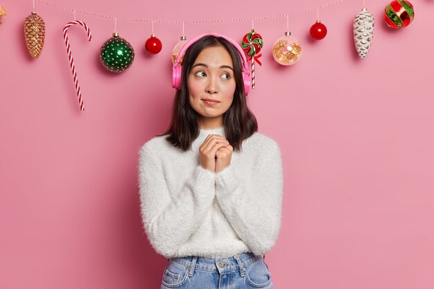 Thoughtful millennial girl keeps hands together being deep in thoughts while listening lyrics song in headphones recalls pleasant memories wears warm winter sweater poses 