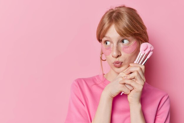 Thoughtful millennial girl with freckled skin keeps lips folded holds cosmetic brushes near face looks away applies beauty patches under eyes isolated over pink bacground copy space for text