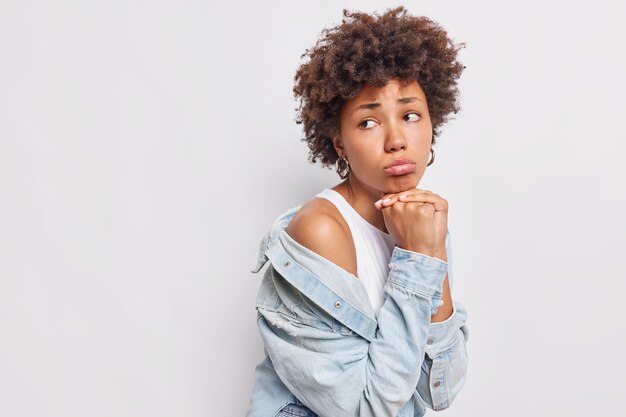 Thoughtful miserable dejected curly Afro American woman keeps hands under chin purses lips looks sadly away wears stylish outfit poses against white wall