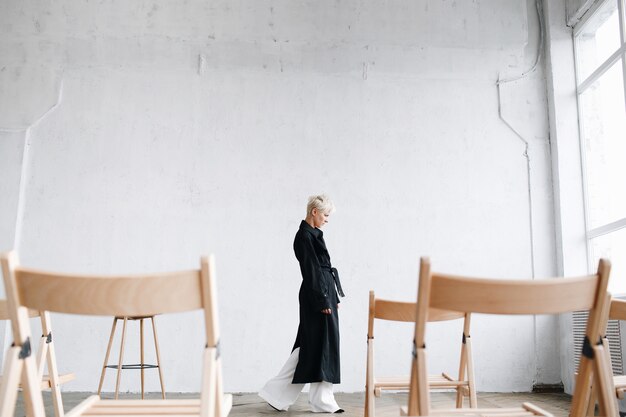 黒いコートの思慮深いモデルがスタジオの椅子の間を歩く