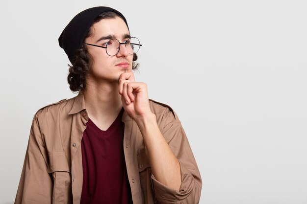 Thoughtful peaceful ambitious hipster looking aside, putting one hand on his chin, posing isolated on light grey, looks attentive and concentrated. Copyspace for advertisement.