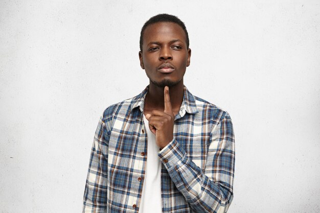 Thoughtful pensive young African American man wearing stylish checkered shirt over white t-shirt keep index finger at his chin while trying to recollect something