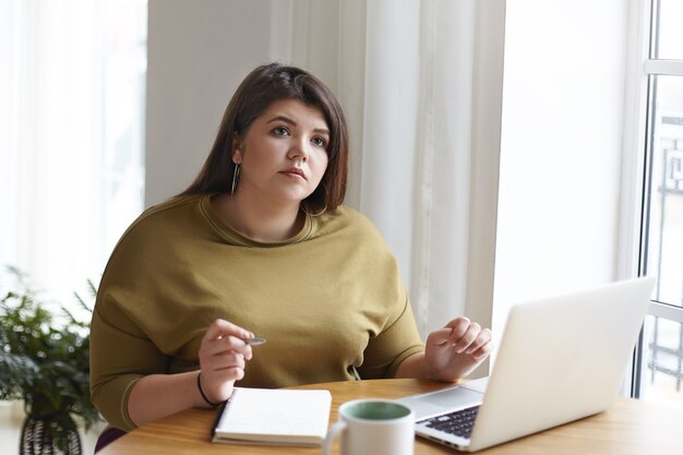 Thoughtful pensive young chubby female journalist sitting at desk with portable computer, mug, looking up while making notes in copybook, working distantly, writing new article for online magazine