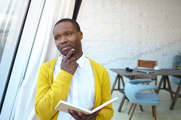 Free photo thoughtful pensive young dark skinned businessman touching chin, standing in office interior, looking through window, planning day, holding diary. people, business, job and occupation concept